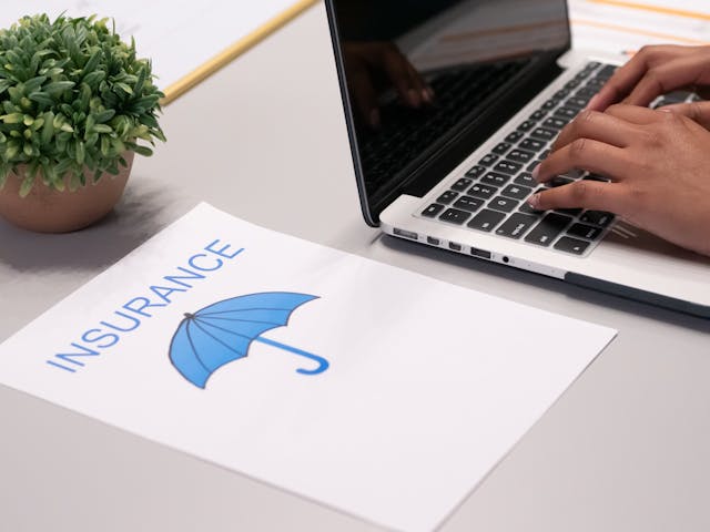 person typing on a laptop next to a sheet of paper that says INSURANCE with a drawing of a blue umbrella below it